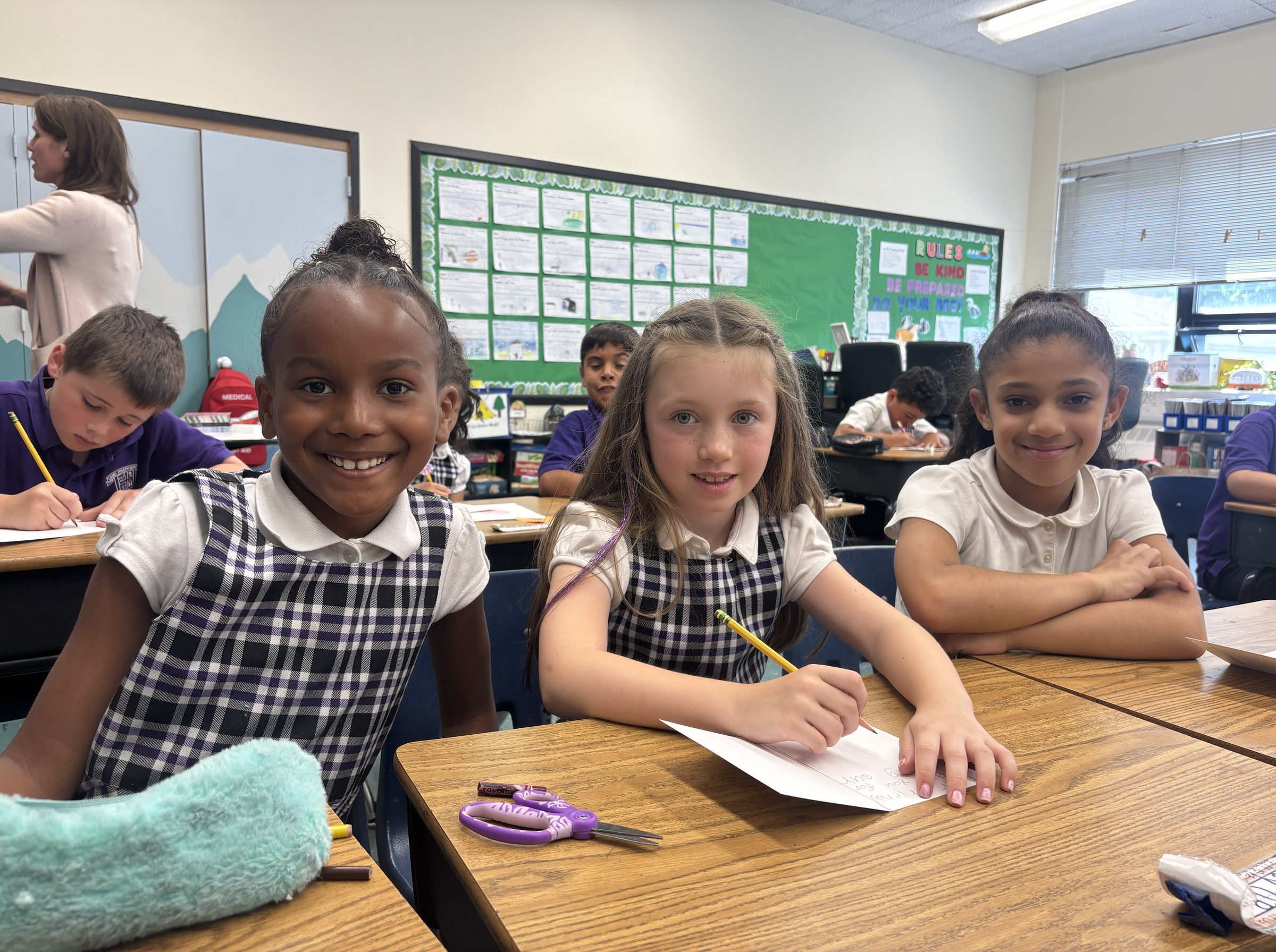Three students in uniform smiling at their desks in a Catholic school classroom, one of them writing with a pencil